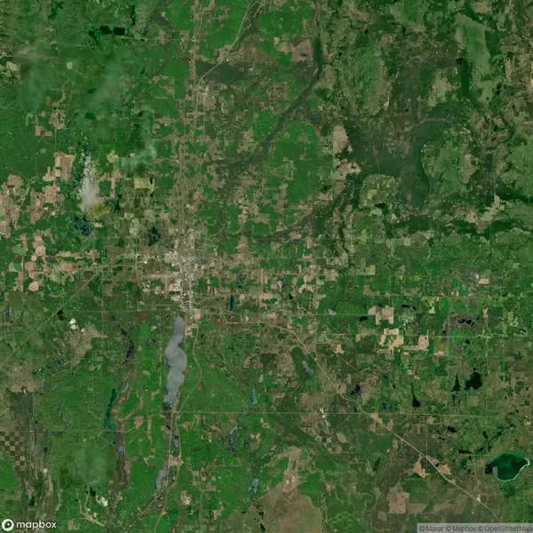 Satellite image showing a mix of urban development, farmland, forests, and lakes in a predominantly green landscape. Roads and fields are visible throughout the area.
