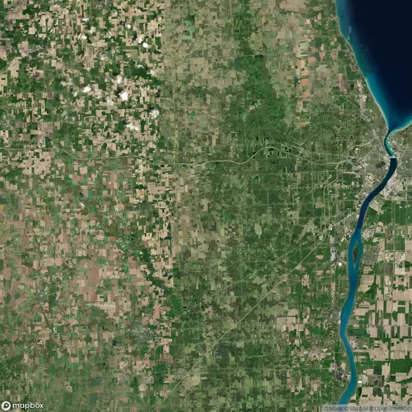 Satellite image showing a patchwork of farmland, a river running vertically on the right, and a city near the water’s edge. The landscape is mostly green with some brown fields.