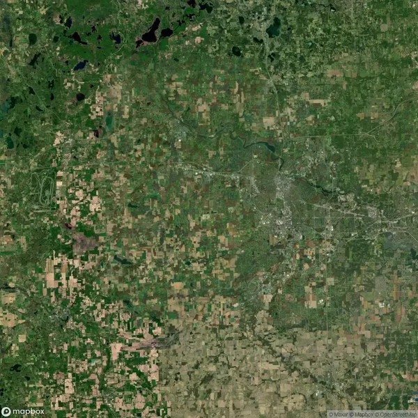 Satellite view of a patchwork landscape featuring agricultural fields, scattered forested areas, and a visible city with a network of roads and waterways.