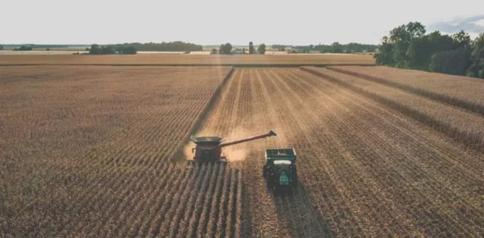 A combine harvester unloads crops into a tractor trailer in a large, sunlit field during harvest.
