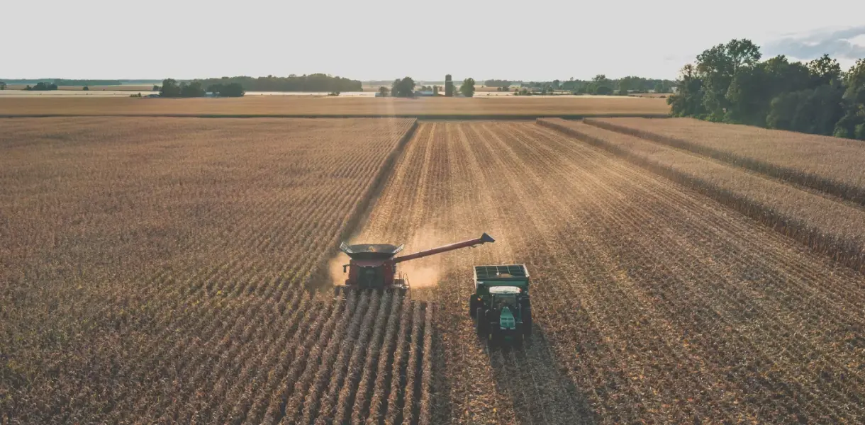 A combine harvester unloads crops into a tractor trailer in a large, sunlit field during harvest.
